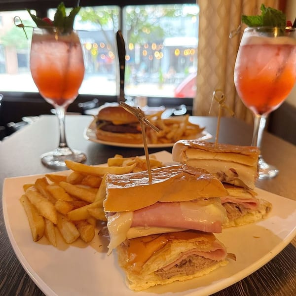 Cuban sandwiches and fries on a white plate, with two red cocktails and a burger in the background.