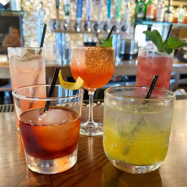 Five colorful Cuban cocktails with garnishes on a wooden bar, with blurred bottles in the background.